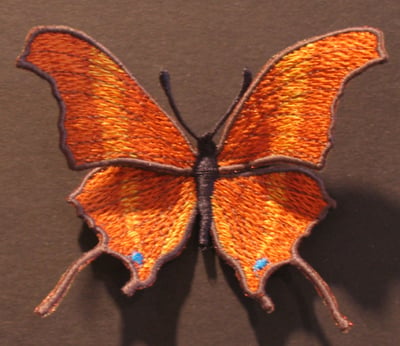 Embroidered orange butterfly with black outlines on brown fabric, blue spots near the hindwings.