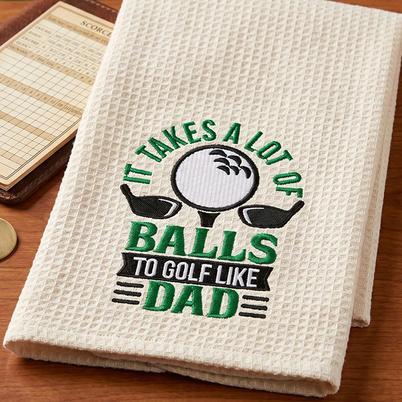 lotofball_GolfTowel | Embroidery Legacy White textured towel with green and black embroidery reading 'It takes a lot of balls to golf like Dad,' featuring a golf ball and two clubs.