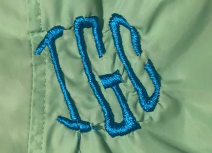 Blue satin-stitch monogram embroidered on lightweight fabric, showing significant puckering and fabric distortion around the letters.