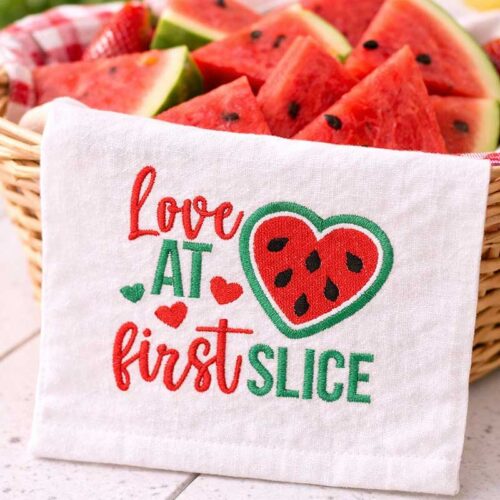 Decorative towel with the phrase 'Love at first slice' and a watermelon heart, beside a basket of watermelon slices.
