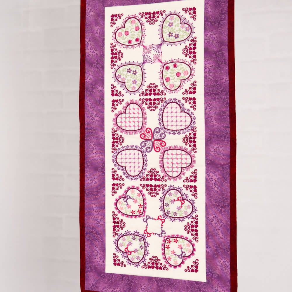Artboard26 Decorative quilted wall hanging with a white center panel of pink hearts and floral designs, framed by purple borders.