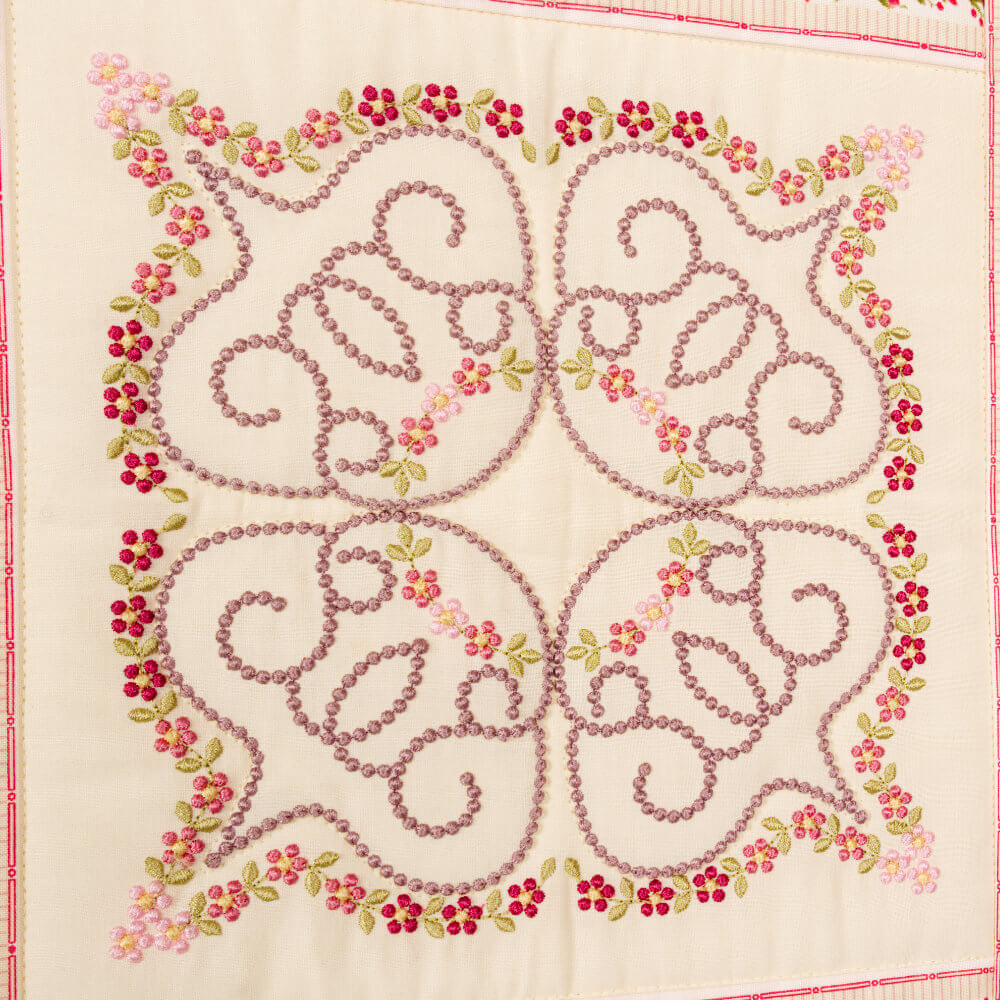 Decorative embroidery on cream fabric with lavender looping scrolls and pink floral borders.
