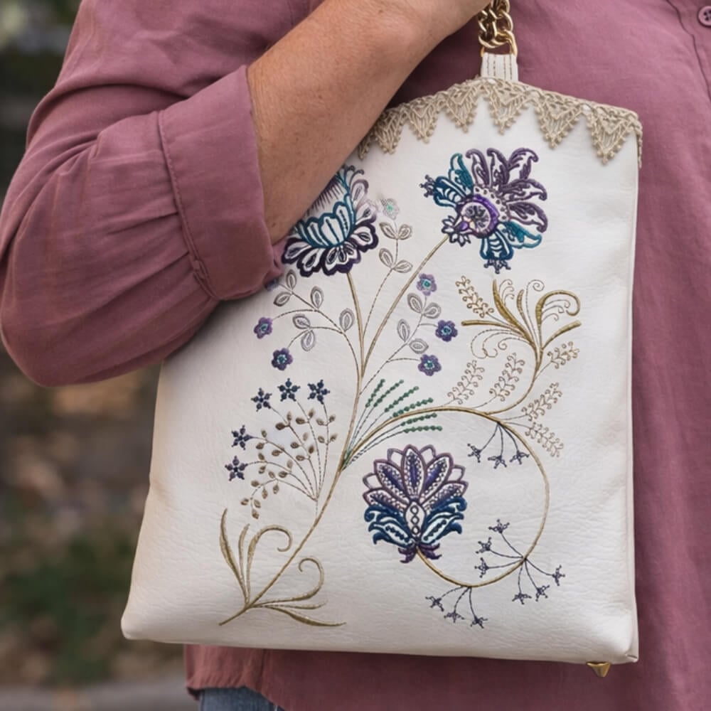 White fabric tote bag with colorful floral embroidery being held by a person wearing a mauve shirt袖