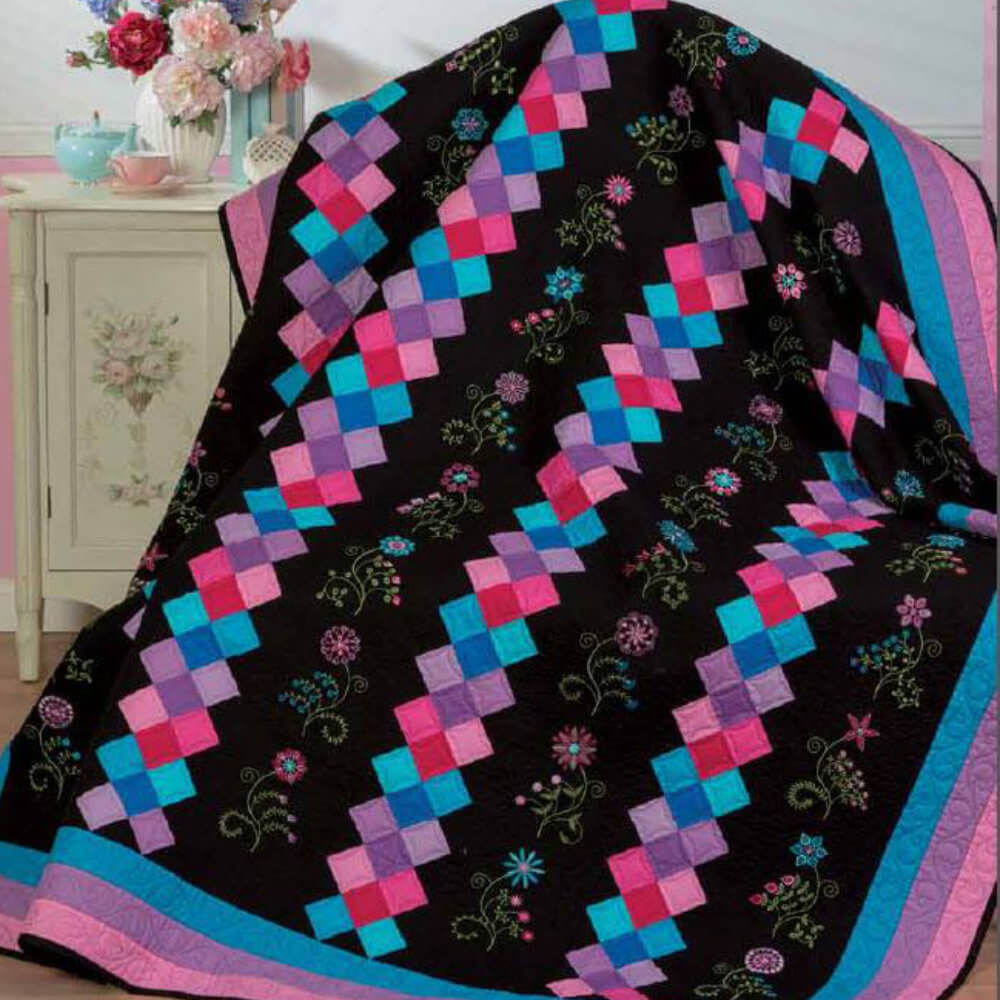 Artboard46 Patchwork quilt draped over a chair/sofa: black background with pink, purple, and blue squares plus embroidered florals on the borders.