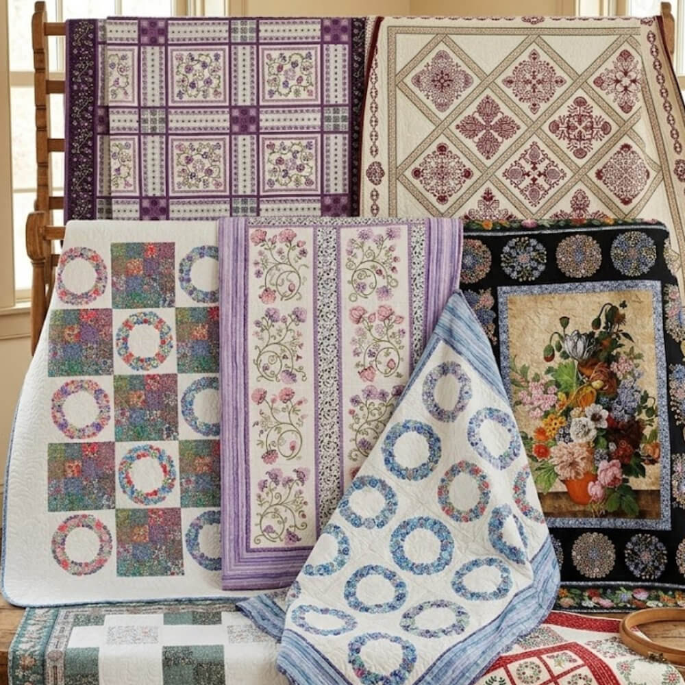 Artboard47 A display of colorful quilts draped over a rack, featuring floral and geometric patterns in purple, lavender, blue and cream.