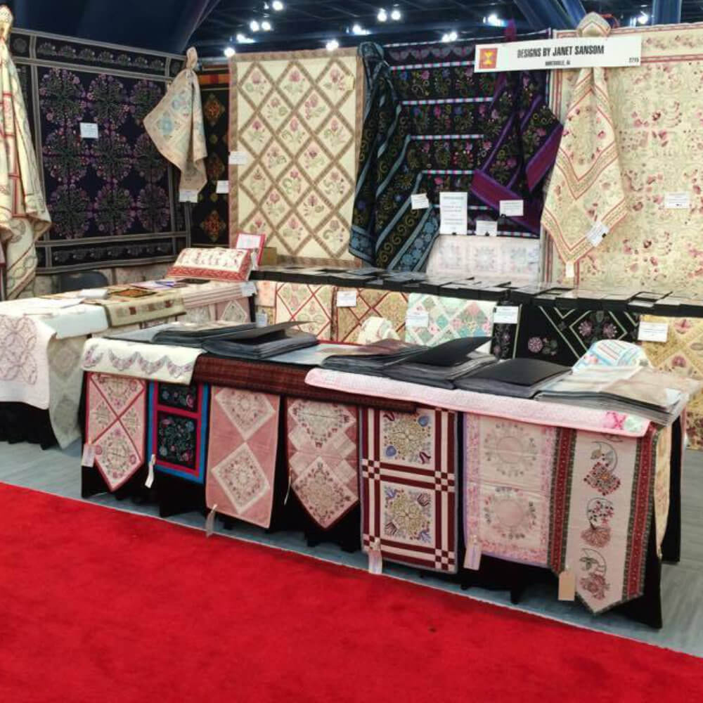 Artboard47_2 Booth at a textile fair displaying patchwork quilts and embroidered fabrics hung on walls and folded textiles laid out on tables over a red carpet