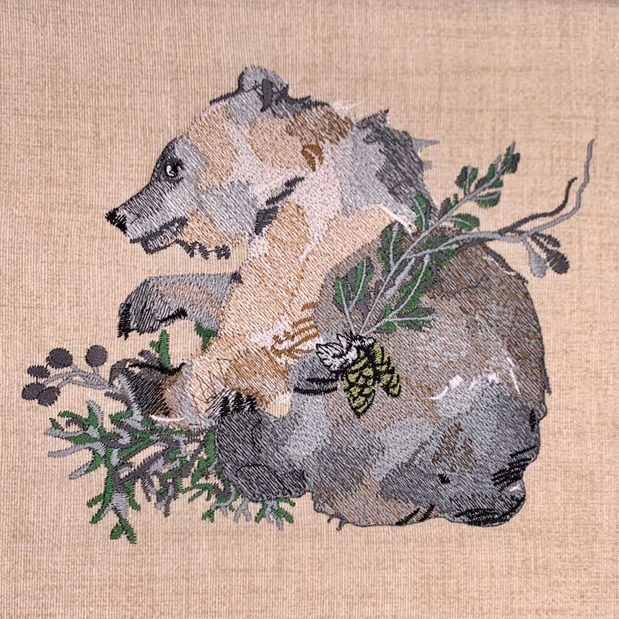 Embroidered artwork of a tan and gray dog seated with green leaves and a pinecone motif on beige fabric