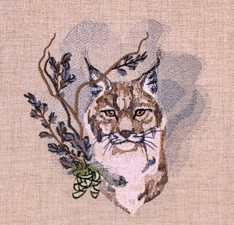 Embroidered image of a lynx head with branches and leaves on a beige fabric background, in brown and tan tones.