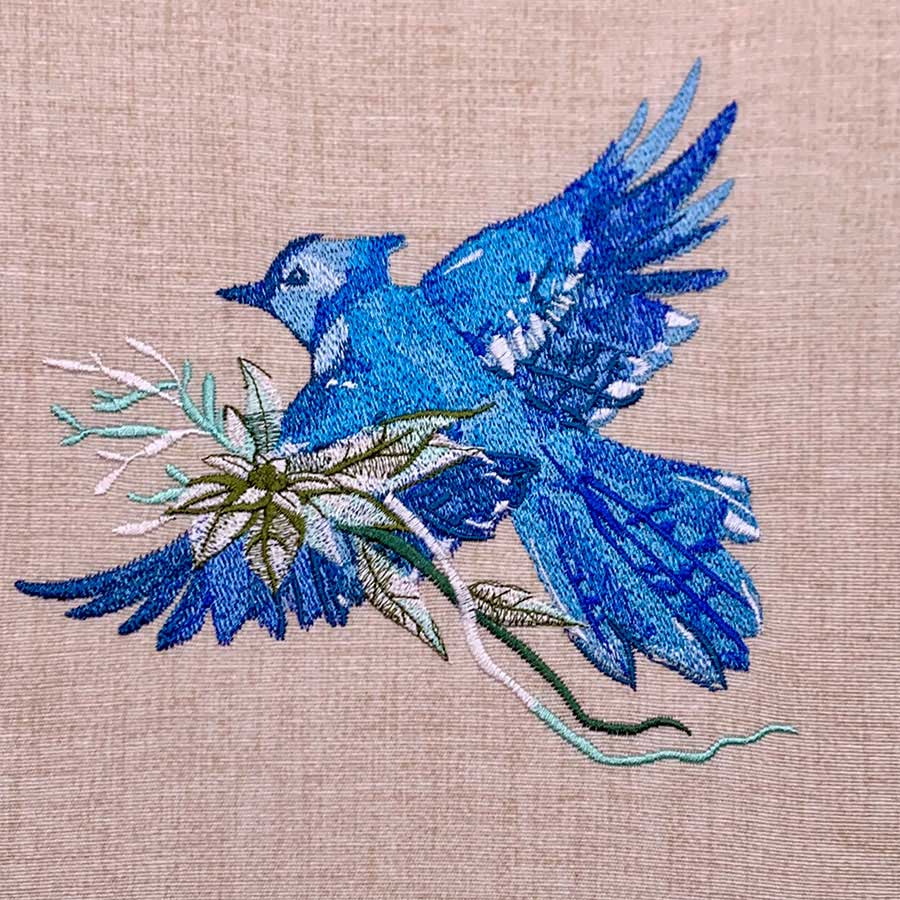 Decorative embroidery of a vivid blue bird in flight on beige fabric, gripping a small branch with green leaves.