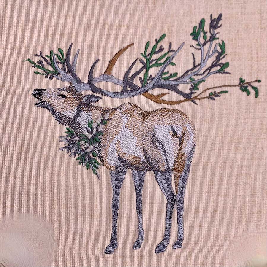 Embroidered illustration of a reindeer with large branching antlers on beige fabric with greenery around its neck
