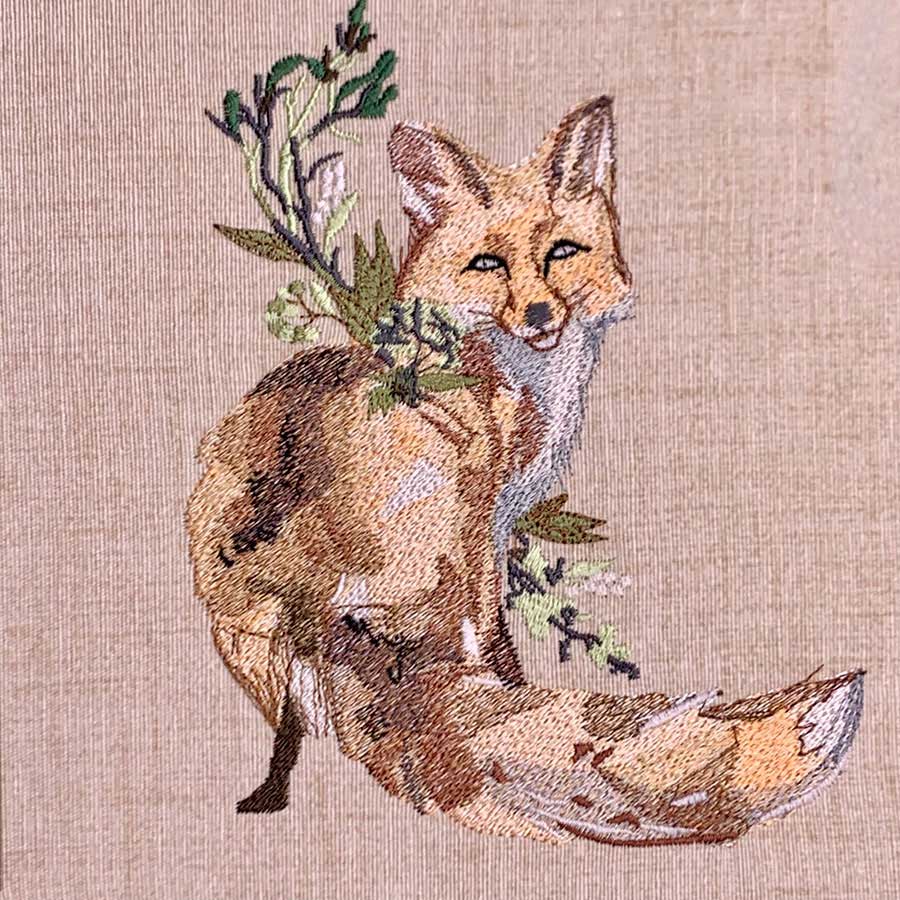 Embroidered fox sitting with green sprigs on its back against beige fabric material, decorative textile art.