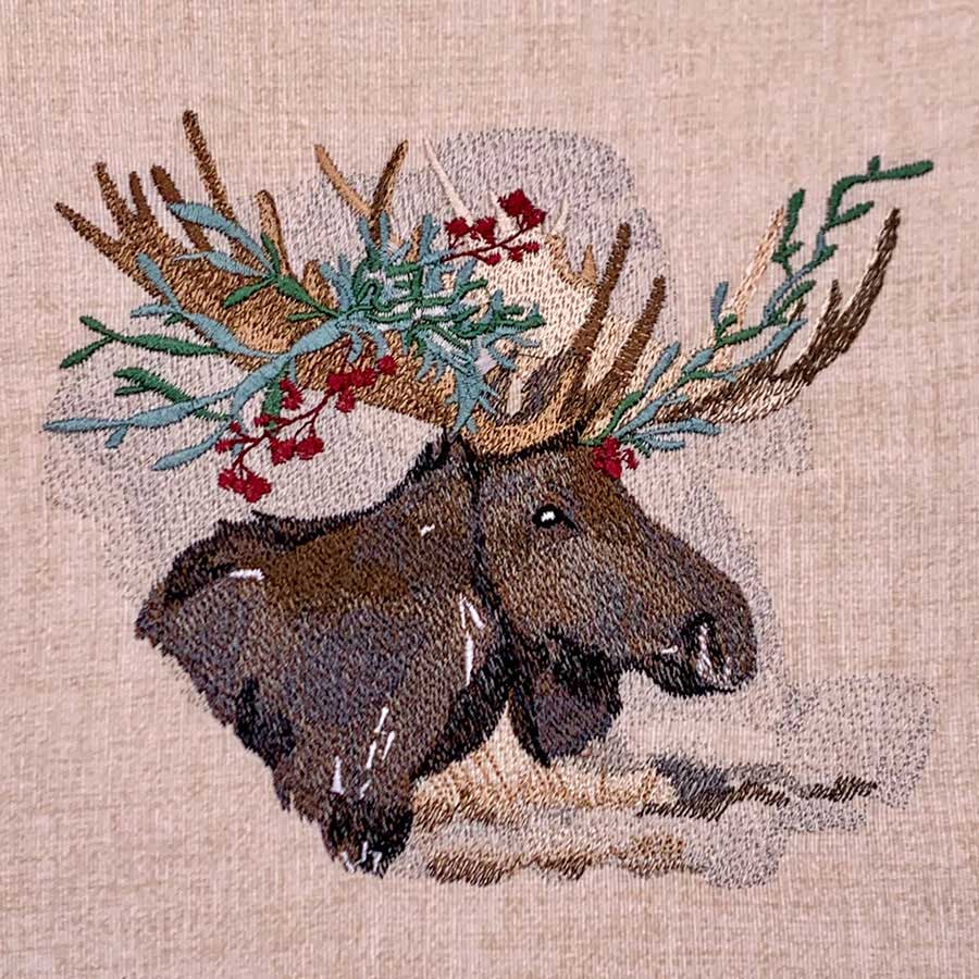 Embroidered tapestry showing two moose heads with antlers adorned with greenery and red berries on a beige background.