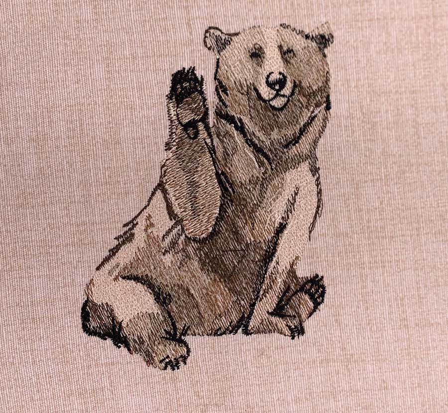Embroidery of a smiling brown bear sitting and waving with its right paw on beige fabric.