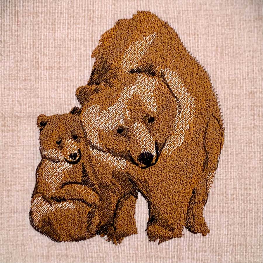 Embroidery-style illustration of a brown bear standing over a cub on a light fabric background.