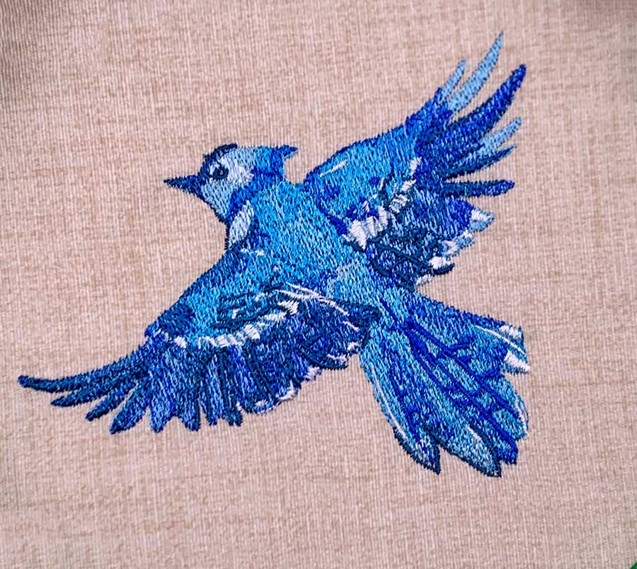 Embroidered blue bird in flight on beige fabric, with spread wings and white highlights in the plumage.