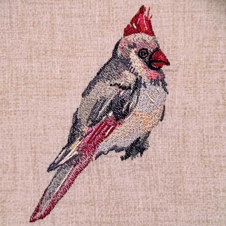 Embroidery of a small cardinal-like bird with red crest and tail on beige fabric.