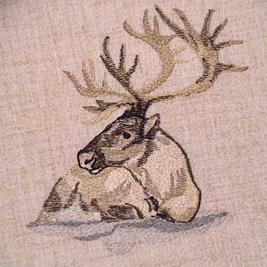 Tapestry-style embroidery of a resting elk with large antlers in the snow on beige fabric
