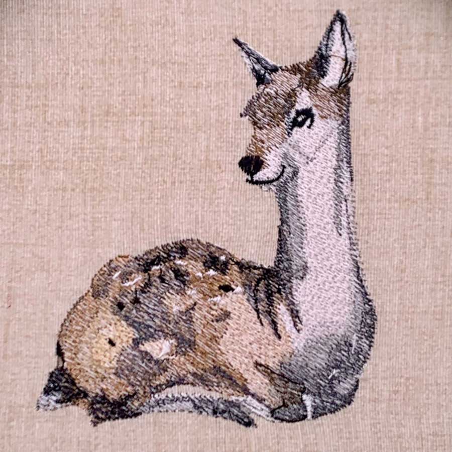 Embroidered illustration of a relaxed llama sitting, facing slightly to the left against a beige background.