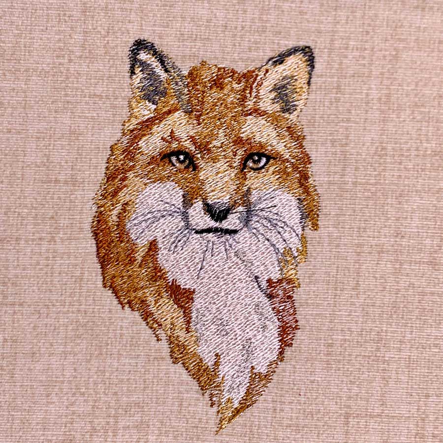 Embroidery of a fox's head stitched in orange, white, and black threads on beige fabric (decorative textile art).