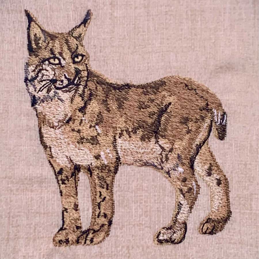 Embroidery of a standing wild cat with tufted ears on beige fabric.