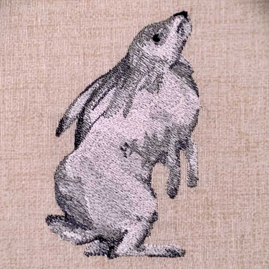 Embroidery of a gray rabbit standing upright on hind legs on beige fabric.