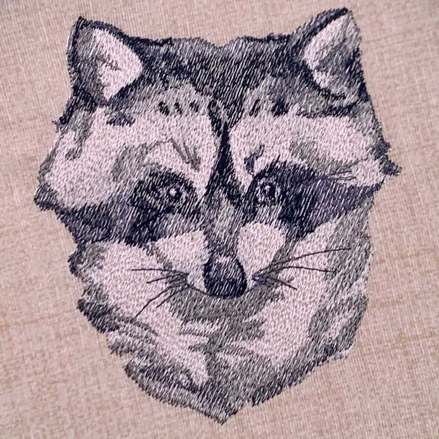 Embroidered cat face design in gray and black on beige fabric.