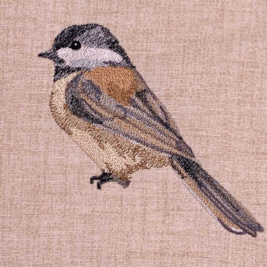 Embroidery of a small brown and gray bird perched on a beige fabric background.