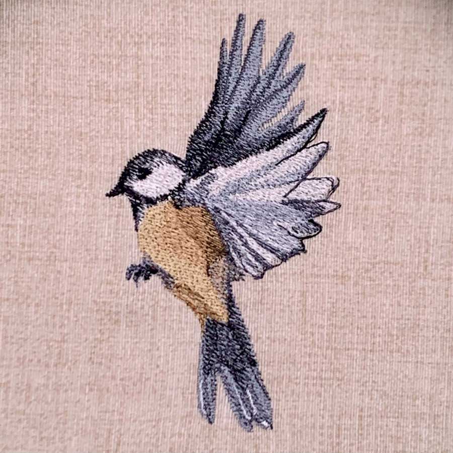 Embroidery of a small bird in flight with white head, orange chest, and gray wings on beige fabric.