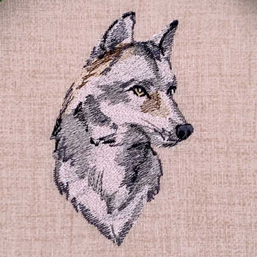 Profile view of a gray wolf's head embroidered on beige fabric, with yellow eye and detailed stitching