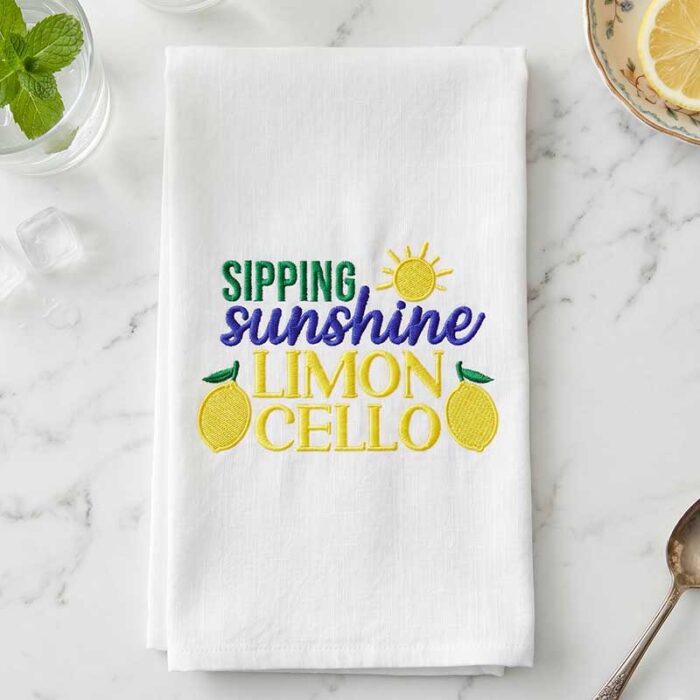 SDMS_SippingSunshine_Napkin | Embroidery Legacy White towel with colorful embroidery reading 'SIPPING sunshine LIMON CELLO' and lemon motifs on a marble surface