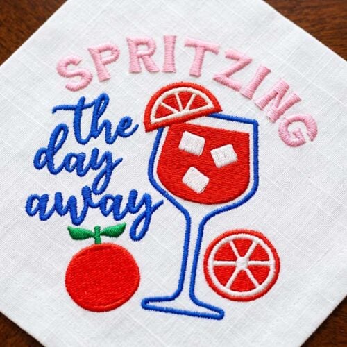 SDMS_SpritzingDay_CocktailNapkin | Embroidery Legacy Embroidered white napkin with a red and blue cocktail glass, orange slice, and the phrase 'SPRITZING the day away' in pink and blue lettering.