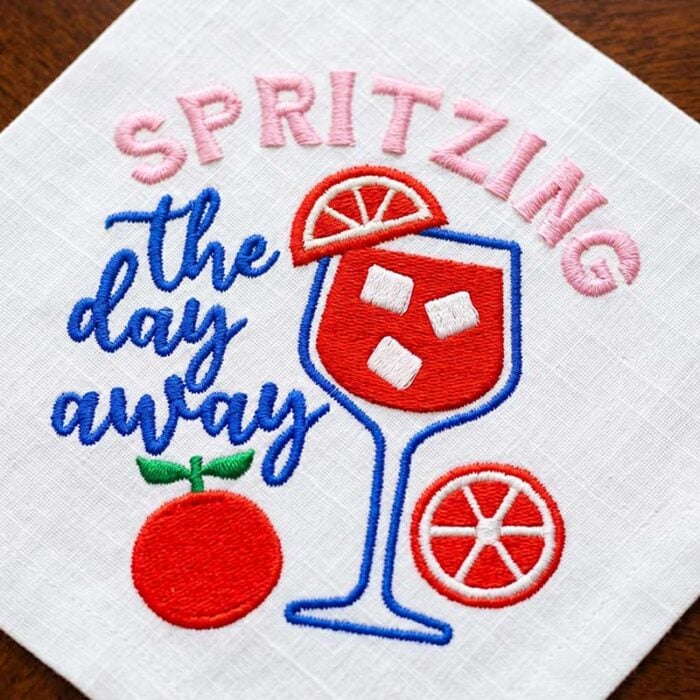SDMS_SpritzingDay_CocktailNapkin | Embroidery Legacy Embroidered white napkin with a red and blue cocktail glass, orange slice, and the phrase 'SPRITZING the day away' in pink and blue lettering.