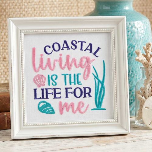 Framed embroidery reading 'Coastal living is the life for me' with pink and teal letters and seashell motifs on a white background.