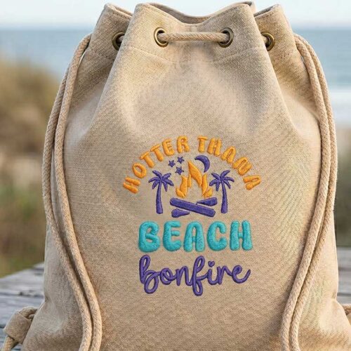 Beige drawstring backpack with embroidered beach bonfire design featuring palm trees and a campfire.
