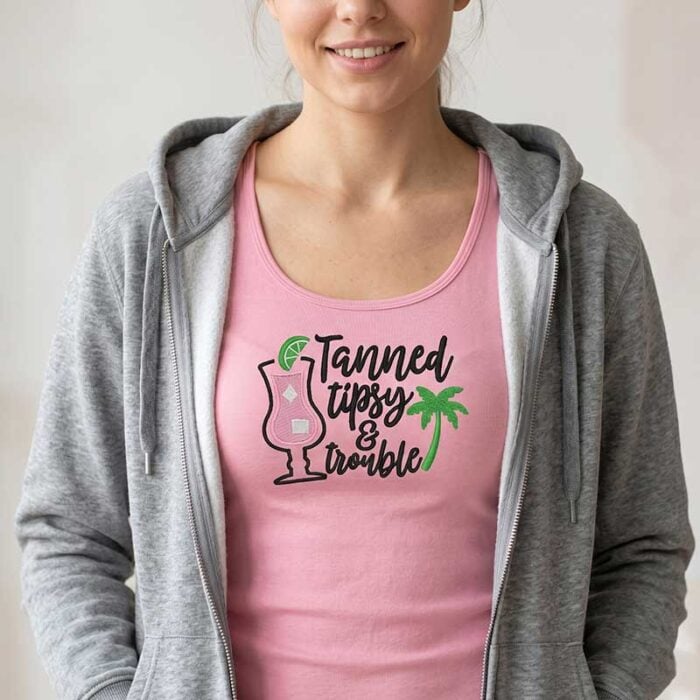 SDSB_TannedTipsy_TShirt2 | Embroidery Legacy Woman wearing a pink tank top with a graphic that says 'Tanned, Tipsy & Trouble' beside a cocktail glass and palm tree, under a gray hoodie