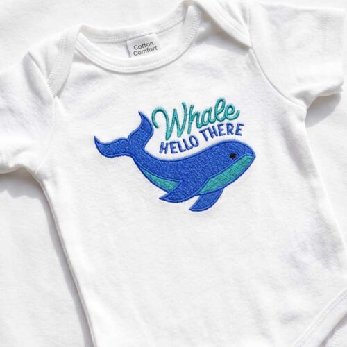 White baby onesie with a blue embroidered whale and the text 'Whale Hello There' on the front.
