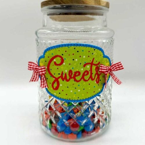 Clear glass jar filled with colorful candy at the bottom; green label says 'Sweets' with blue edge and red gingham ribbons on sides.