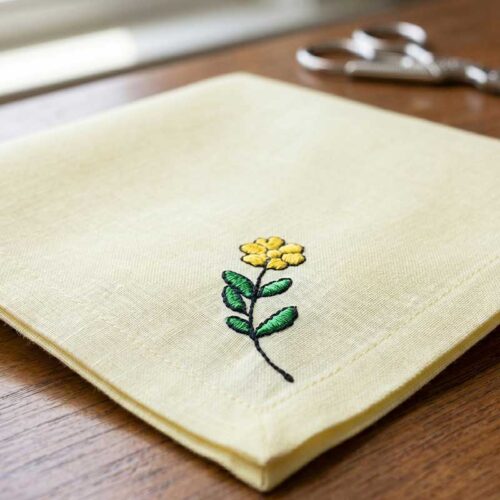 Beige fabric napkin with a yellow embroidered flower and green leaves on a wooden surface; blurred scissors in the background.