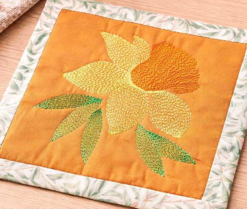 KarenD_FlowerMugRugDownload1 Embroidered orange quilt block with a yellow daffodil and green leaves on an orange center and green patterned border.