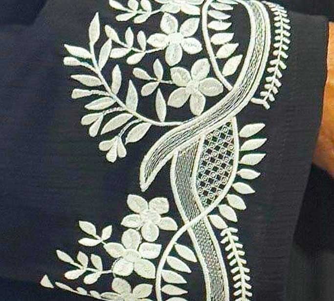MohamedS_Sleeve_Download3 1 Close-up of intricate white floral embroidery on dark fabric, showing a winding vine, leaves, and a lattice-filled leaf motif.