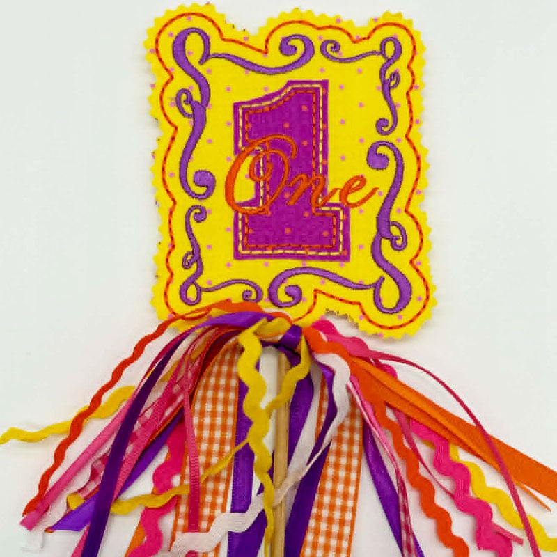 VintageFrames2_Caketopper | Embroidery Legacy Colorful first-birthday banner card with a yellow ornate frame, purple number 1 and the word ‘One,’ and multicolored fringe ribbons cascading below.