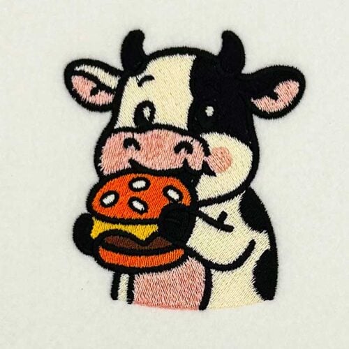 EL_CA_Cow | Embroidery Legacy Embroidered cartoon cow eating a hamburger on a white fabric background and patch edge visible below.