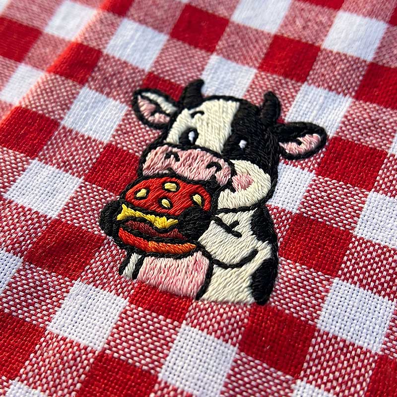 EL_CA_Cow_Napkin | Embroidery Legacy Close-up of a playful cow embroidery on red-and-white checkered fabric.