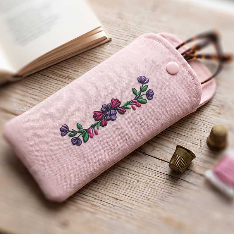 EL_FS4_f_GlassCase | Embroidery Legacy Pink fabric glasses case with purple/pink embroidered flowers on a wooden table, with an open book in the background.