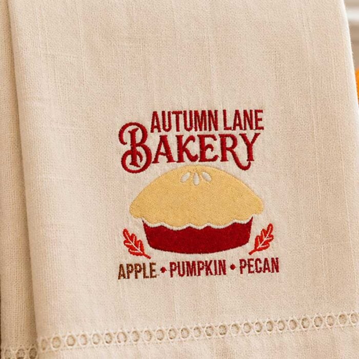 SDFB_AutumnLaneBakery_Towel | Embroidery Legacy Beige embroidered towel showing Autumn Lane Bakery logo with a pie graphic and flavor names: Apple, Pumpkin, Pecan.