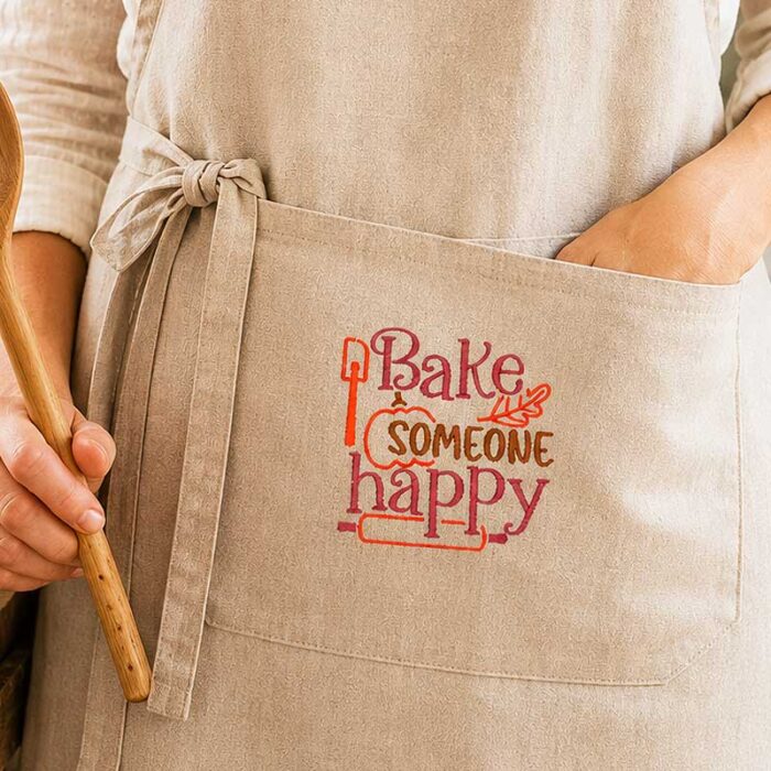 SDFB_BakeSomeoneHappy_Apron | Embroidery Legacy Close-up of a beige apron with an embroidered slogan 'Bake someone happy' and a hand holding a wooden rolling pin in the foreground.