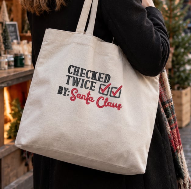 Stop-Searching-Image5 Beige tote bag draped over a person’s shoulder with the text “CHECKED TWICE BY: Santa Claus” and red signature, carried at an outdoor market or store.