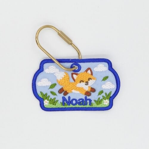 fox-freebie | Embroidery Legacy Embroidered keychain patch with a fox leaping over grass and clouds, the name 'Noah' beneath, bordered in blue with a gold pin attached
