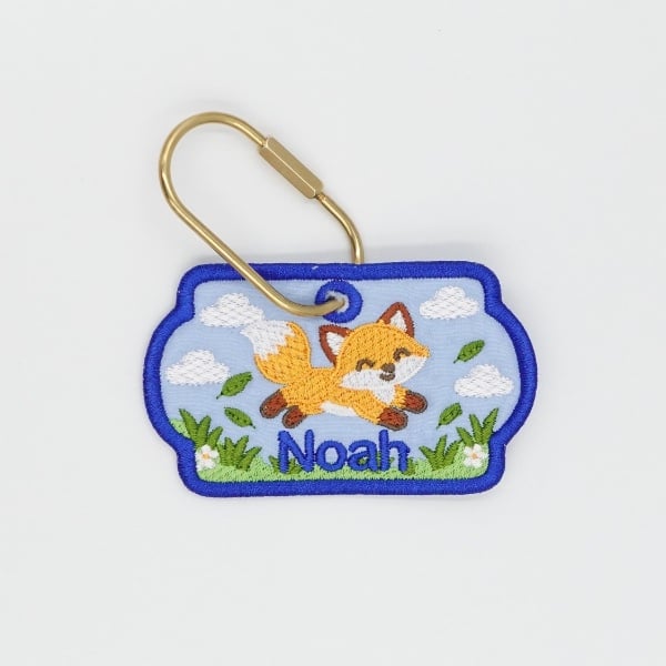 fox-freebie | Embroidery Legacy Embroidered keychain patch with a fox leaping over grass and clouds, the name 'Noah' beneath, bordered in blue with a gold pin attached