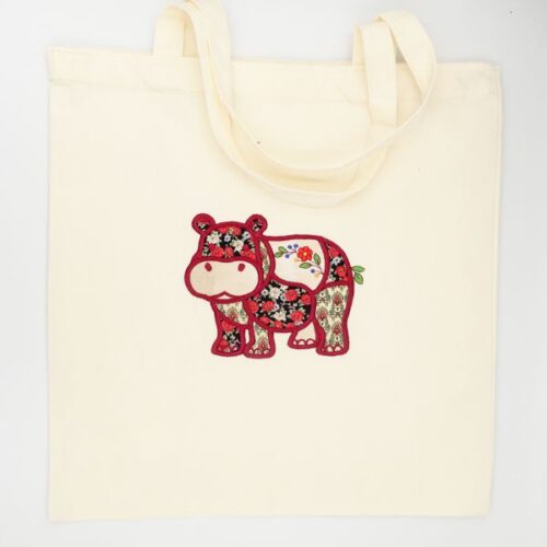 freebie-newsletter-hippo-square | Embroidery Legacy Cream tote bag with a colorful embroidered patchwork hippo design on the front, handles at the top.